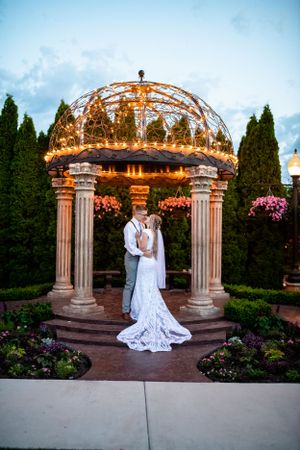 Wedding couple poses under illuminated garden gazebo with stone columns and twinkle lights.