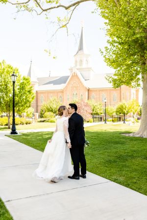 Couple walks along a church sidewalk with green lawn and architecture in background.