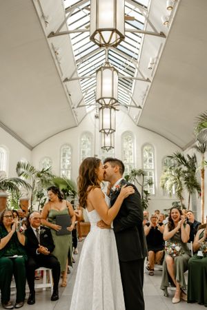 A wedding ceremony kiss in a bright conservatory filled with guests and tropical plants.