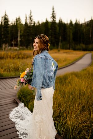 Bride in white dress and denim jacket walks along wooden boardwalk through mountain meadow.