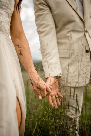 Couple holds hands among tall grass in artistic wedding photo series.