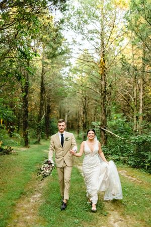 Newlyweds walking along a forest trail surrounded by tall trees and greenery.