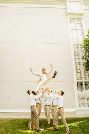 Groomsmen perform playful lifting poses on grass outside a building.
