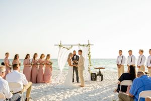 Beach wedding ceremony setup with white chairs and arch against ocean backdrop at sunset.