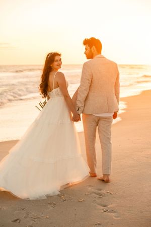 Couple walking along beach at sunset in formal attire leaving footprints in sand.