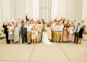 Wedding party dressed in beige and white poses together outside a light colored building.