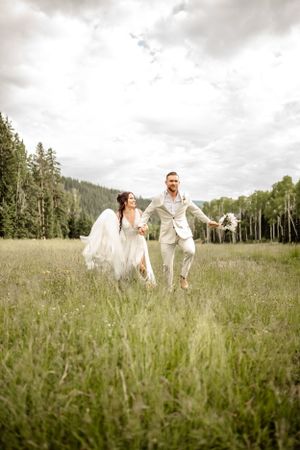 Wedding couple walks through meadow with mountains and forest in background.