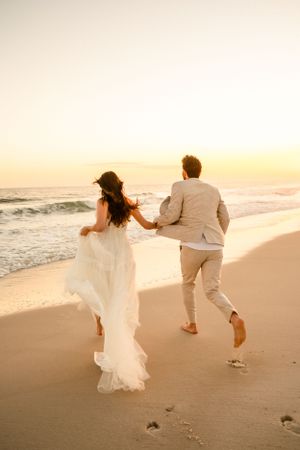 Playful couple runs along beach at sunset in wedding attire.