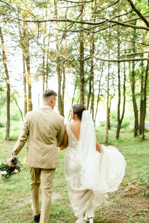 Wedding couple walking through a sunlit forest path with flowing white dress and tan suit.