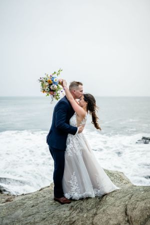 A joyful wedding couple embraces on coastal rocks with crashing waves behind them.
