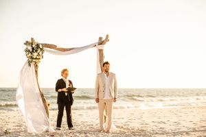 A beach wedding ceremony under a rustic driftwood arch with white fabric and ocean views.