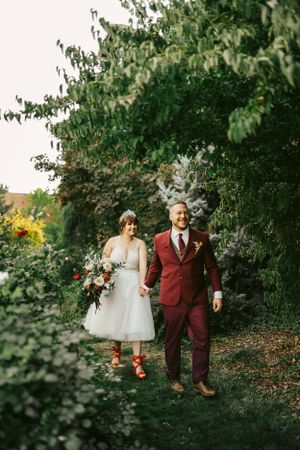 Couple in burgundy and white attire walking through a lush garden path.