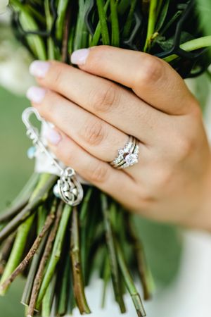 Close up of a sparkling diamond engagement ring and wedding band on a hand holding a bouquet with greenery.
