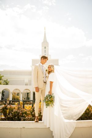 Wedding couple poses outside white temple building with flowing veil in breeze.