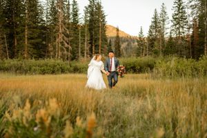 Wedding couple walks through tall grass with mountain peaks and forest in distance.