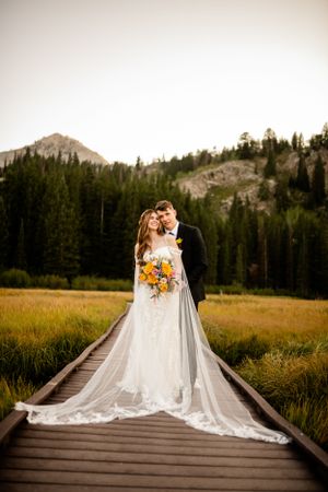 Wedding couple poses on wooden boardwalk with flowing veil and mountain landscape behind them.