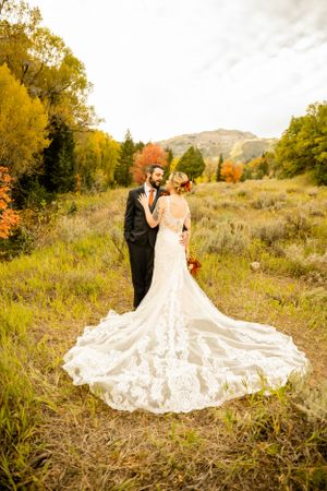 Wedding couple in formal attire standing in autumn field with mountains and dramatic train.