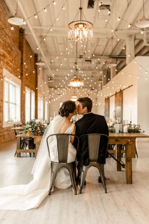 Wedding couple seated in an elegant reception venue with twinkling lights and rustic wooden tables.