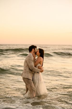 Couple embracing in ocean waves during sunset beach photo session.