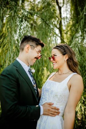 Romantic wedding couple embraces under willow trees in a garden setting.