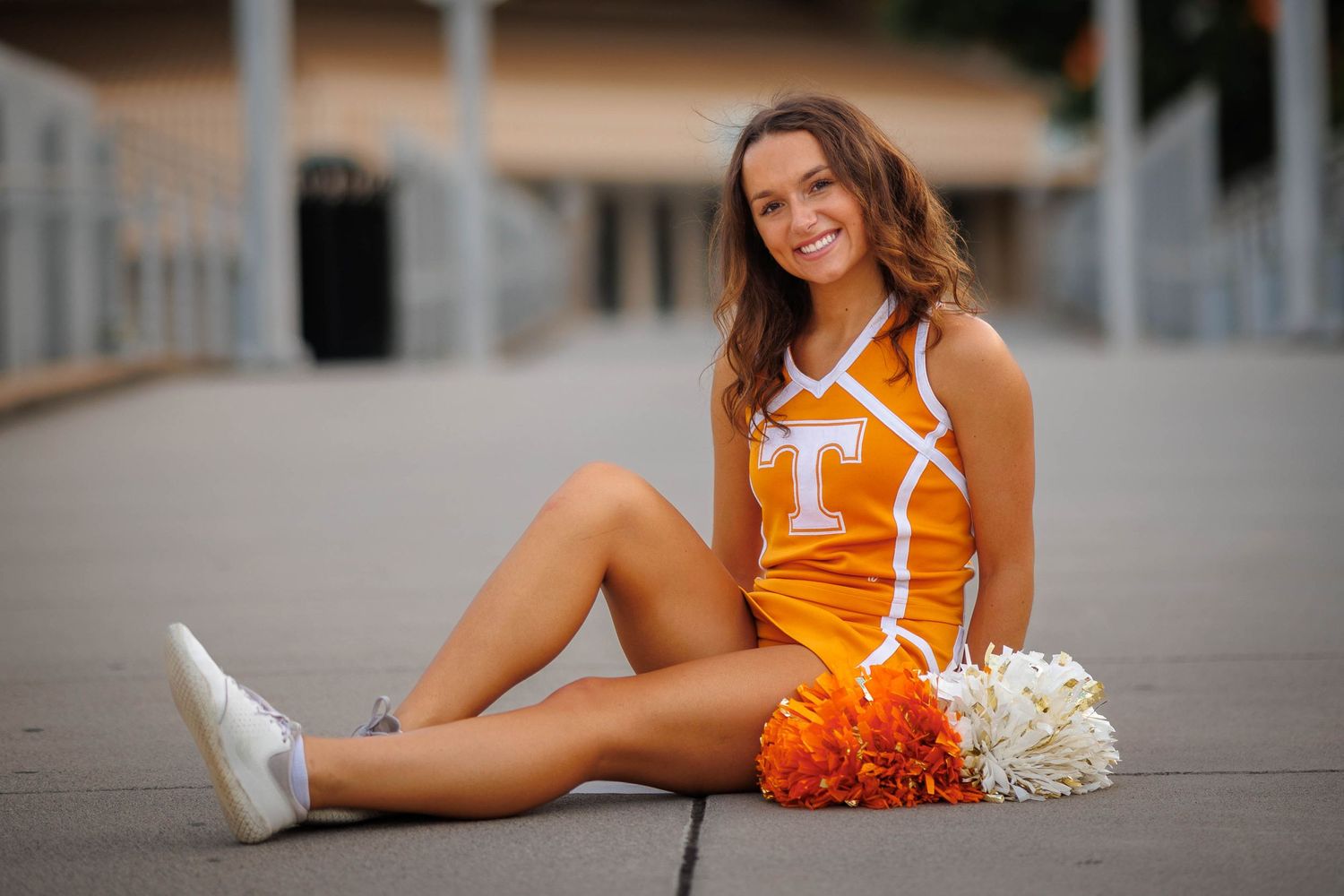 Cheerleader in orange and white uniform sitting with pom poms on concrete.