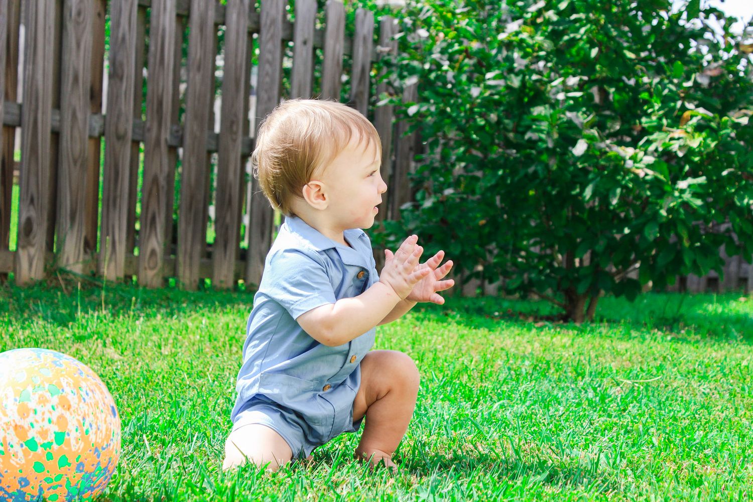 Toddler clapping hands while playing with colorful ball in backyard garden.
