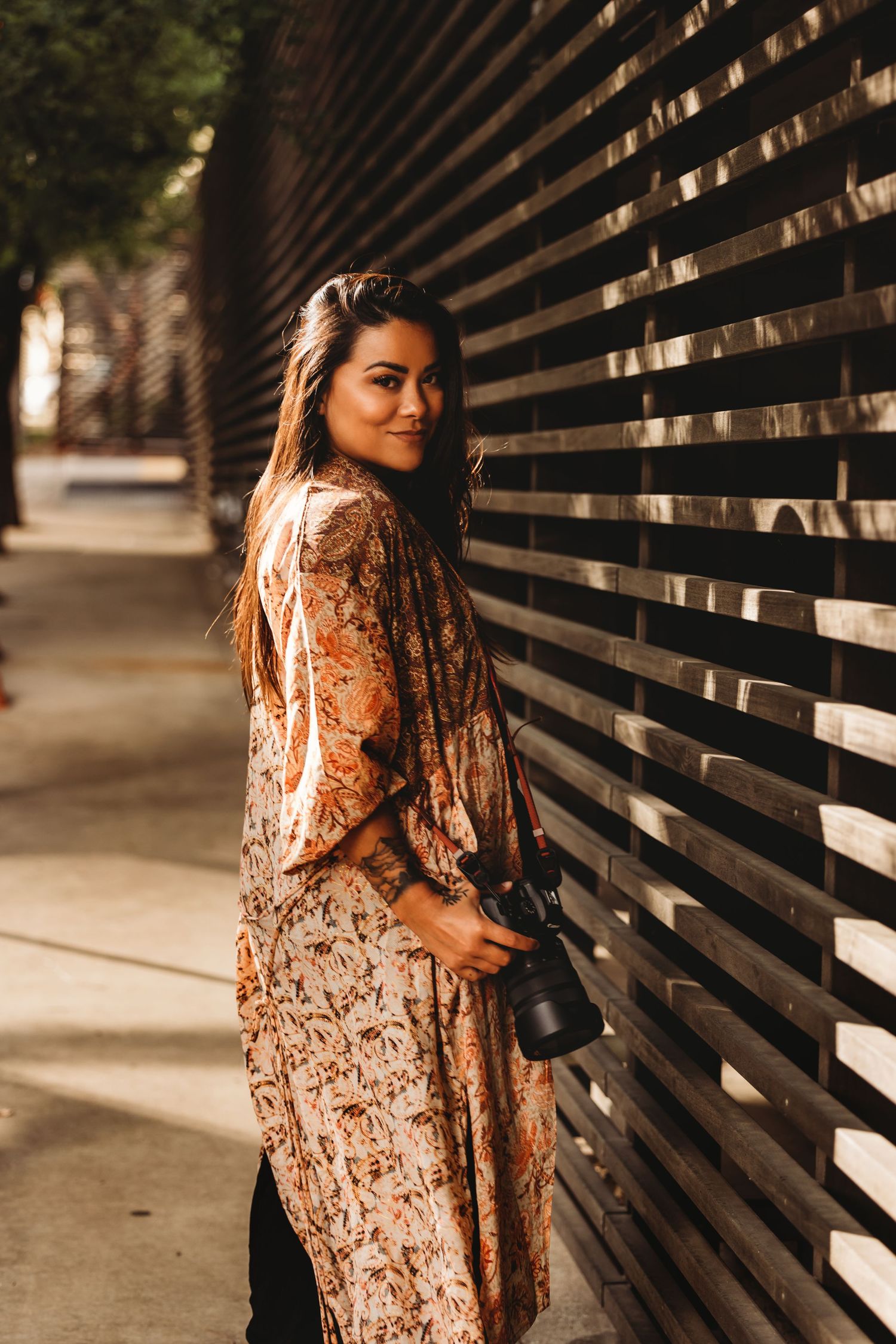 Woman in floral dress poses against wooden slat wall with camera.