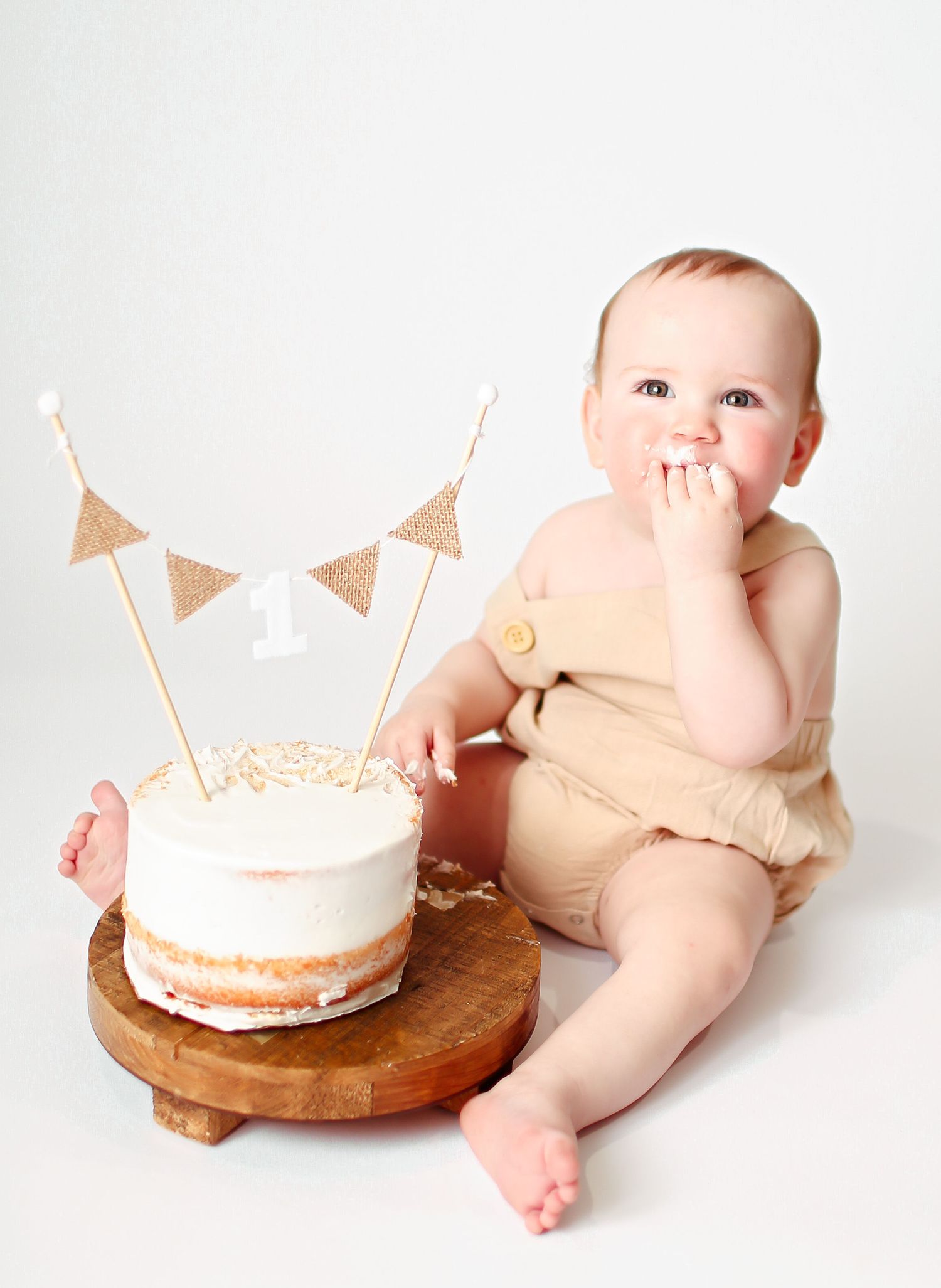 Sweet photo of a baby in neutral clothing beside a simple white cake decorated with burlap bunting flags on wooden stand.