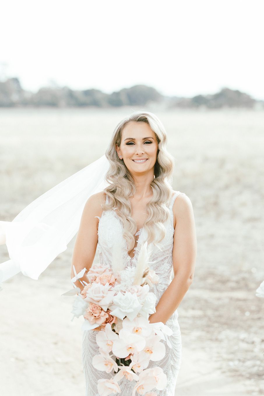 Bride in a lace wedding dress holding a pink bouquet against a bright outdoor backdrop.