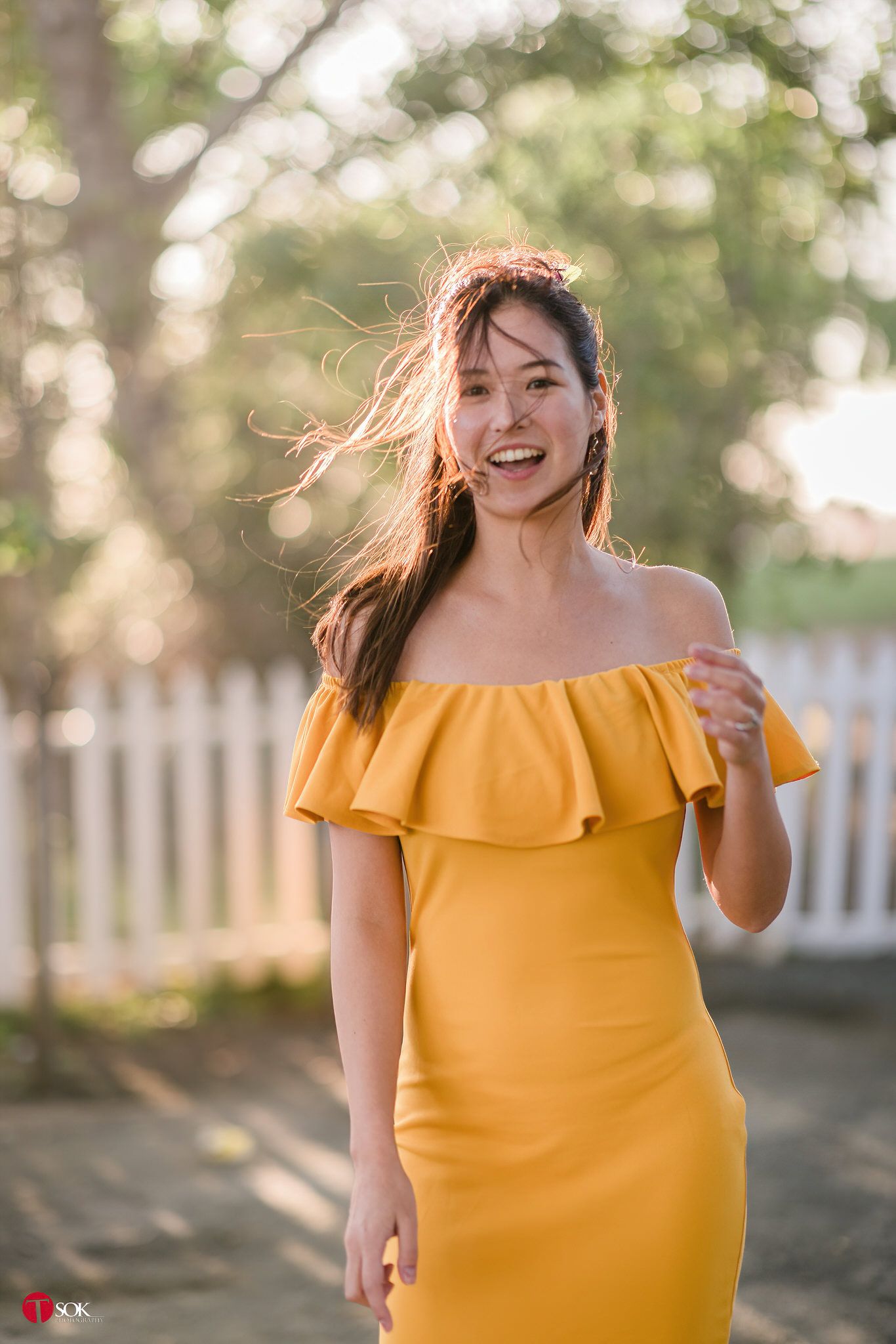 A woman in a yellow dress smiles joyfully outdoors with wind-blown hair.