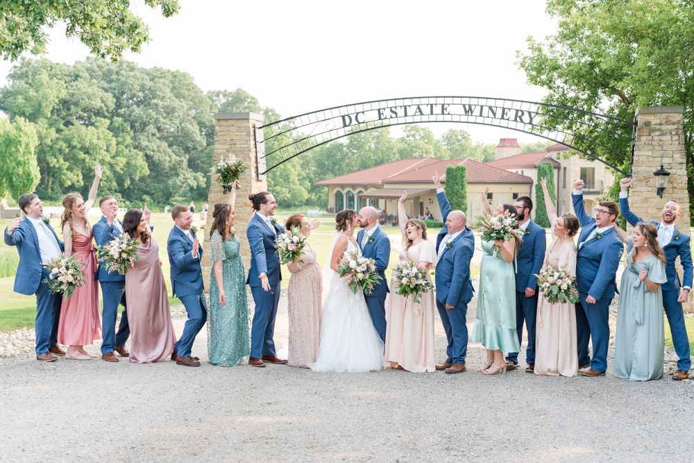 Wedding party in pastel colors poses outdoors at an estate winery with green trees in the background.