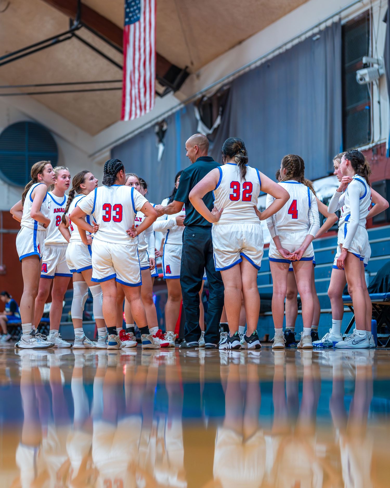 Basketball team huddles with coach on sideline during game with American flag visible overhead.