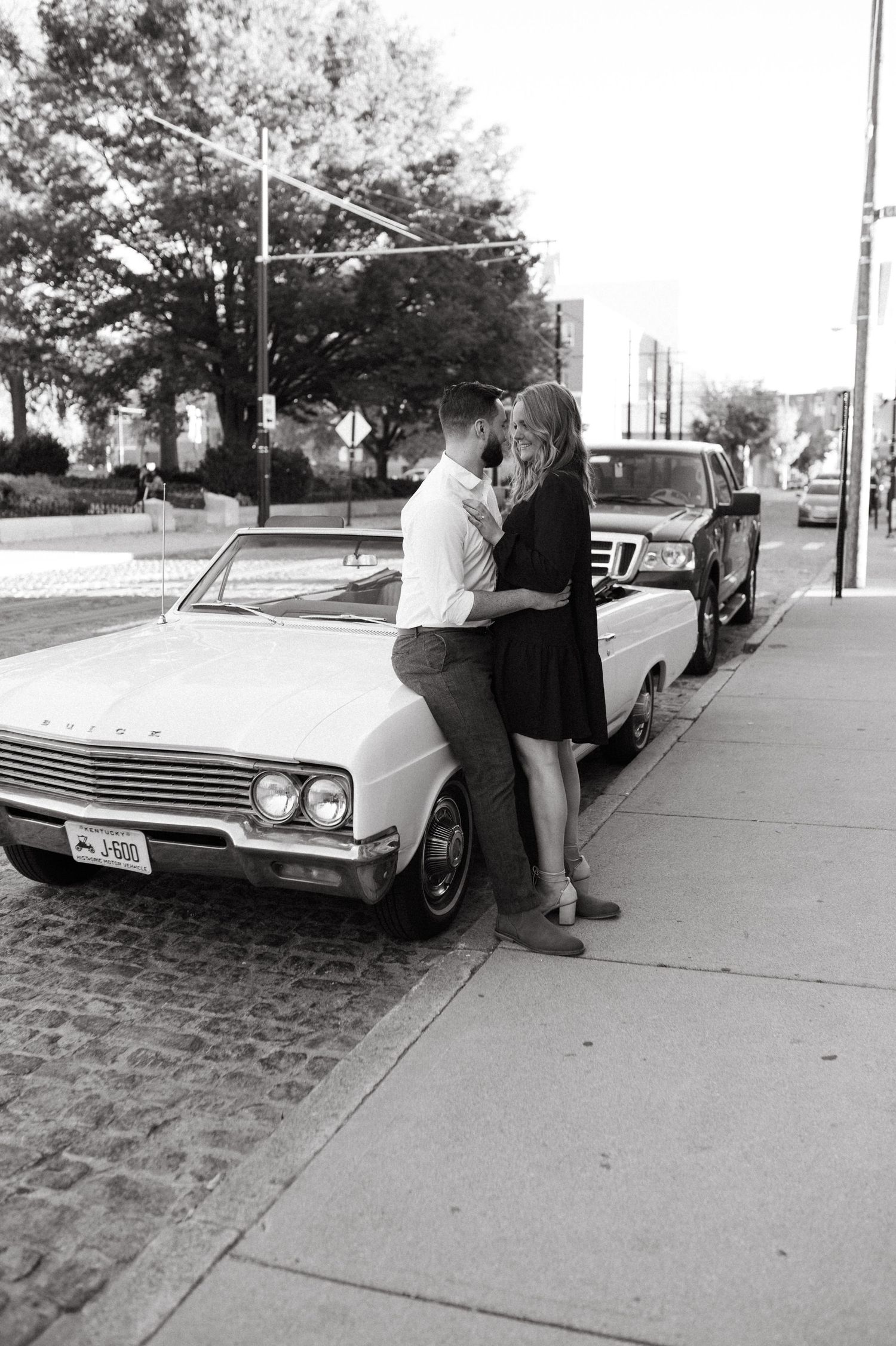 A vintage-style black and white photo of a couple posing by their classic car.