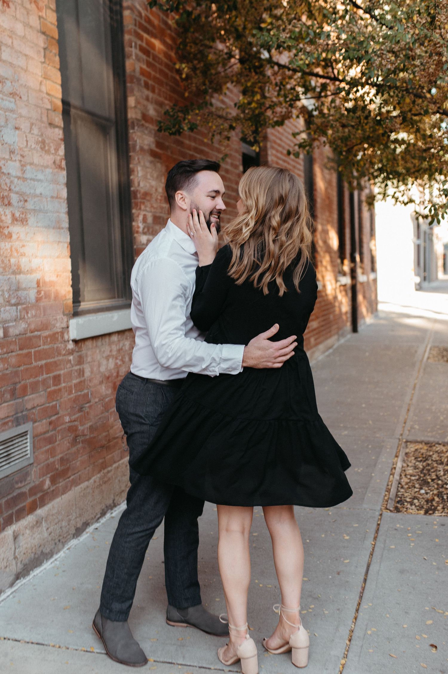 A romantic couple embraces on a brick-lined sidewalk, the woman in a black dress and the man in a white shirt.