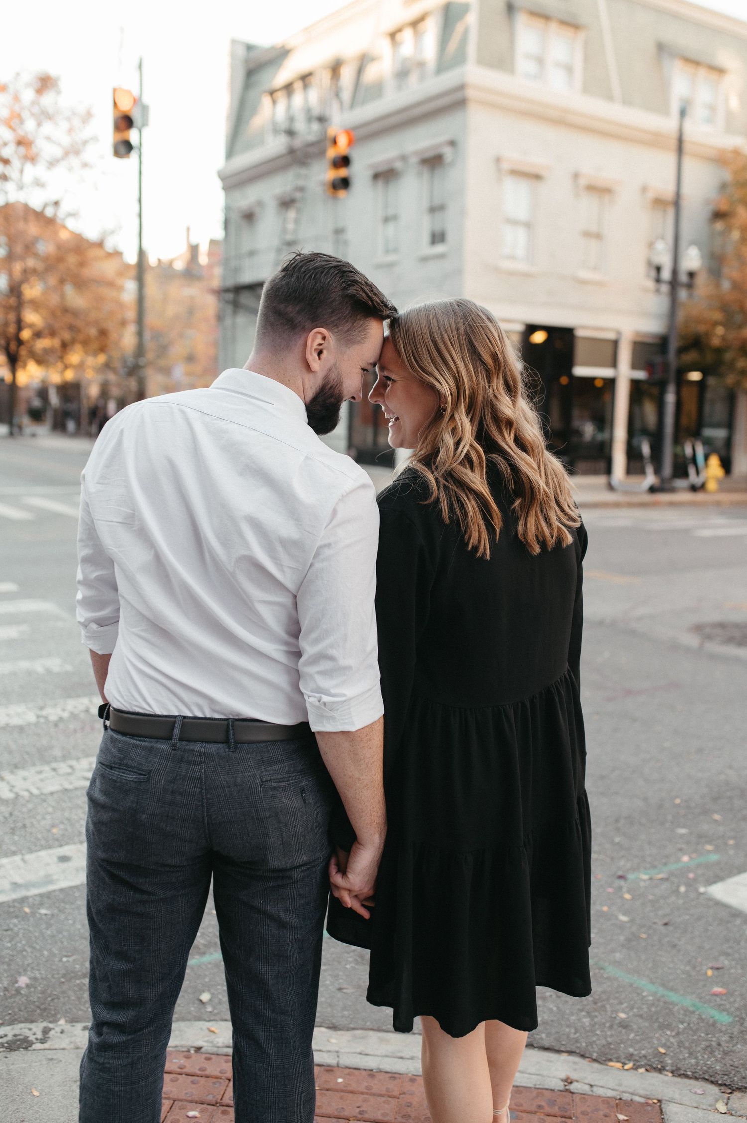 Couple sharing a tender moment on a downtown street corner with autumn trees in the background.