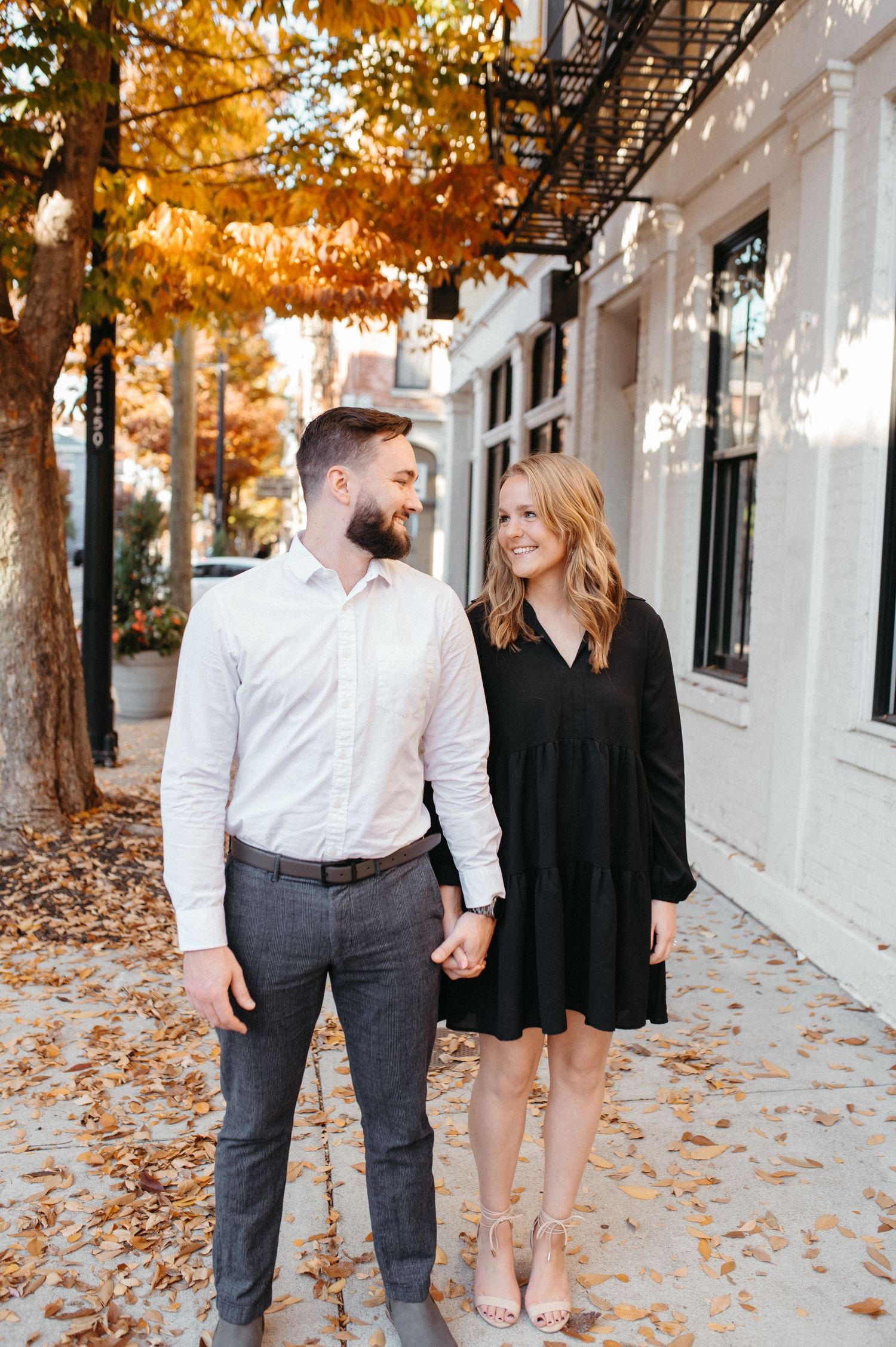 Couple walking hand in hand on leaf-covered sidewalk during fall.