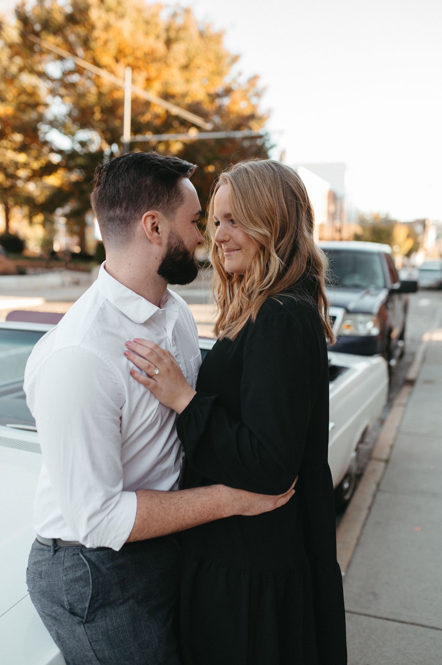 A tender embrace between a couple dressed in black and white beside a parked vintage car.