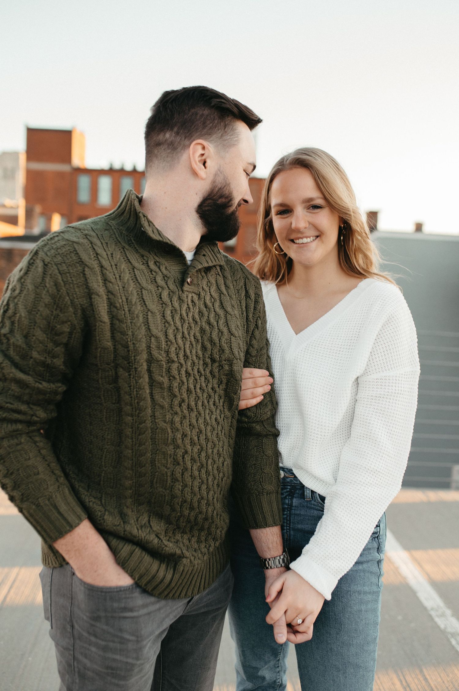 A couple in casual sweaters embraces on a city rooftop during sunset.