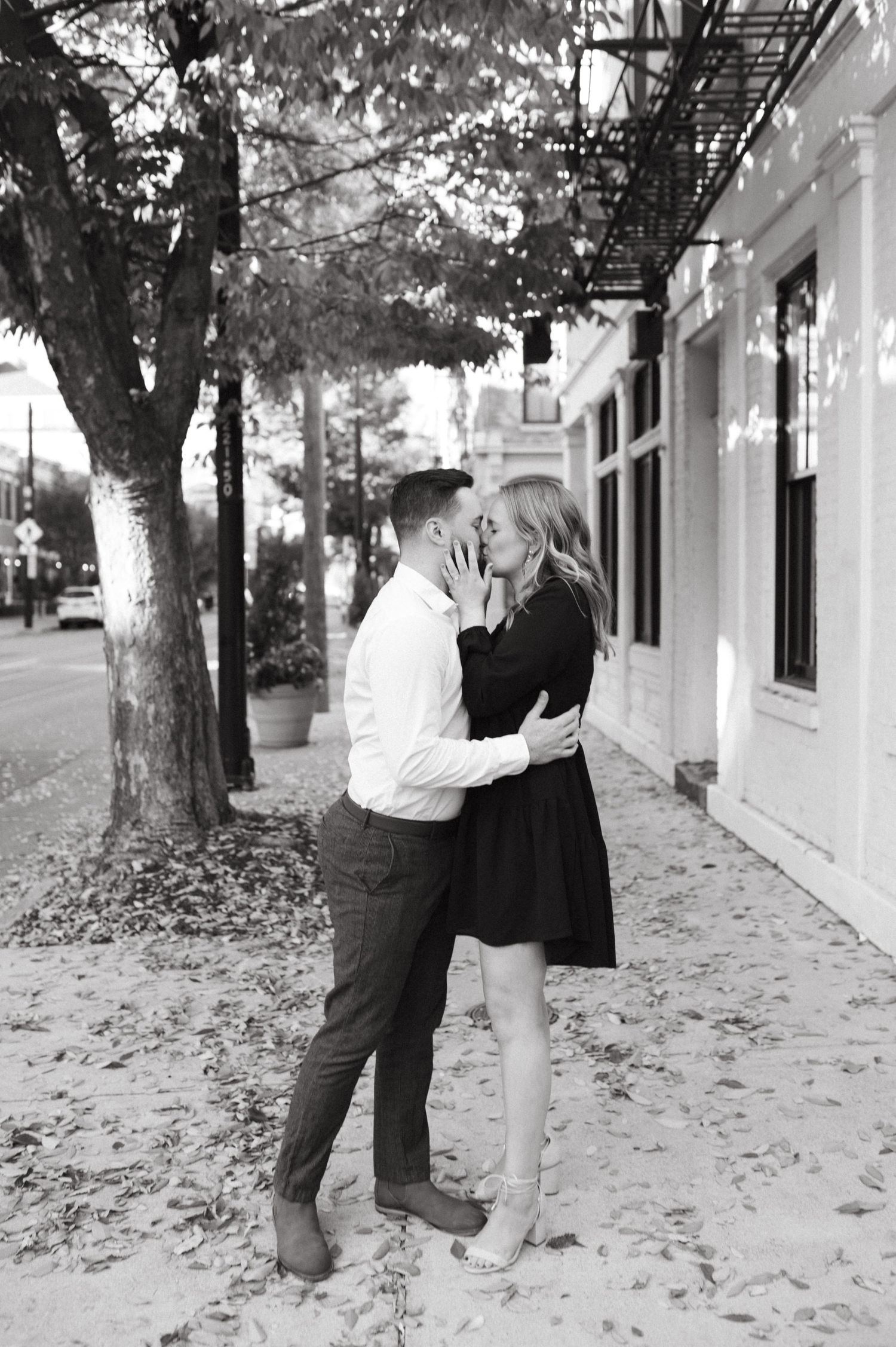 A romantic black and white portrait of a couple embracing on a tree-lined sidewalk next to a white building.