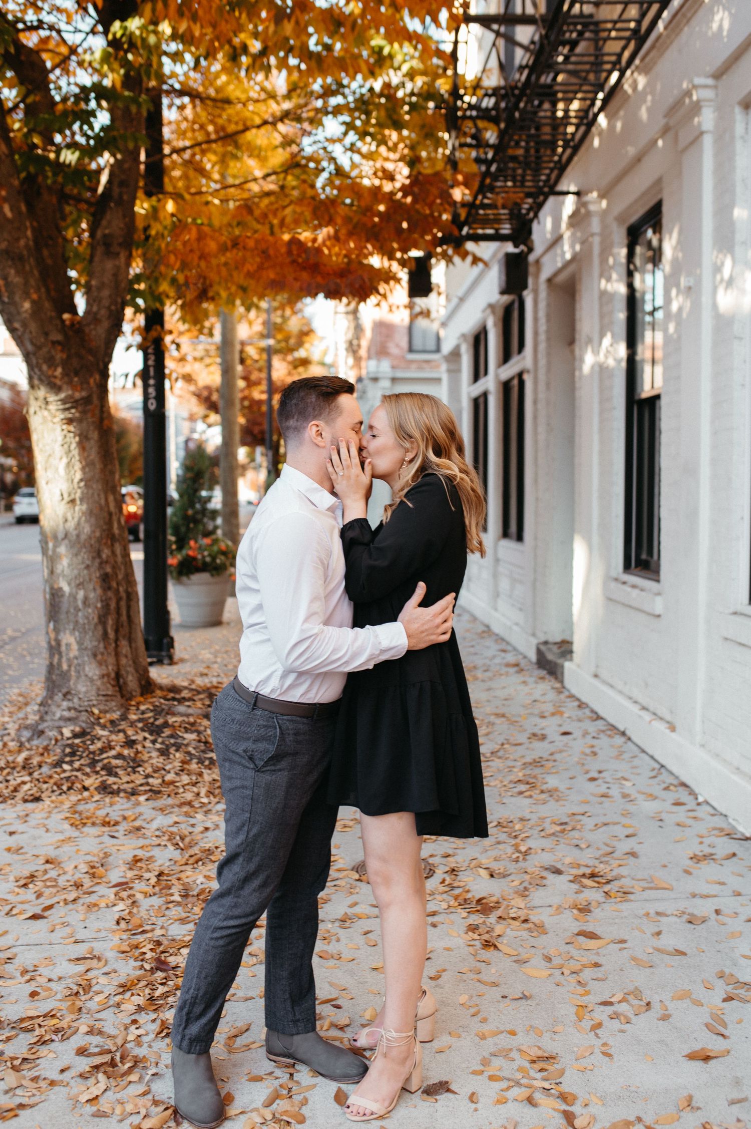 A couple sharing an intimate moment under autumn trees with orange leaves falling near a white storefront.