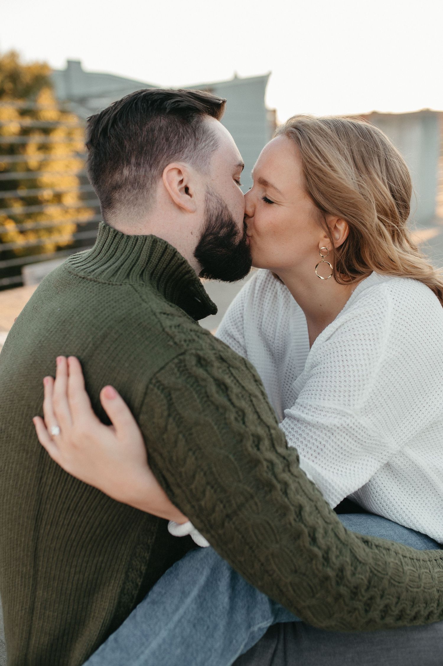 Romantic couple sharing a tender kiss while wearing cozy sweaters on a rooftop at sunset.