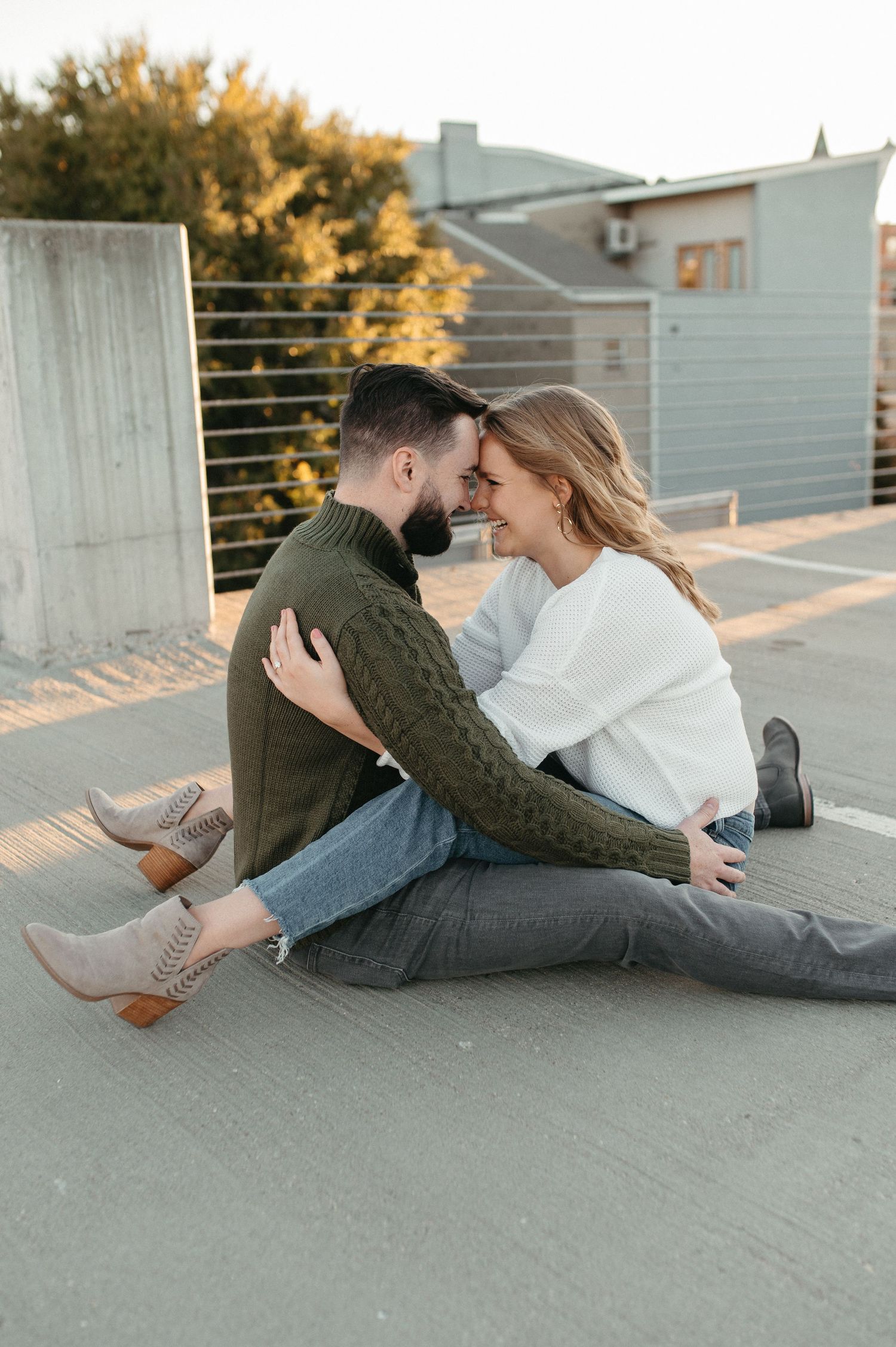 A romantic couple in knit sweaters embrace while sitting on a rooftop at sunset.