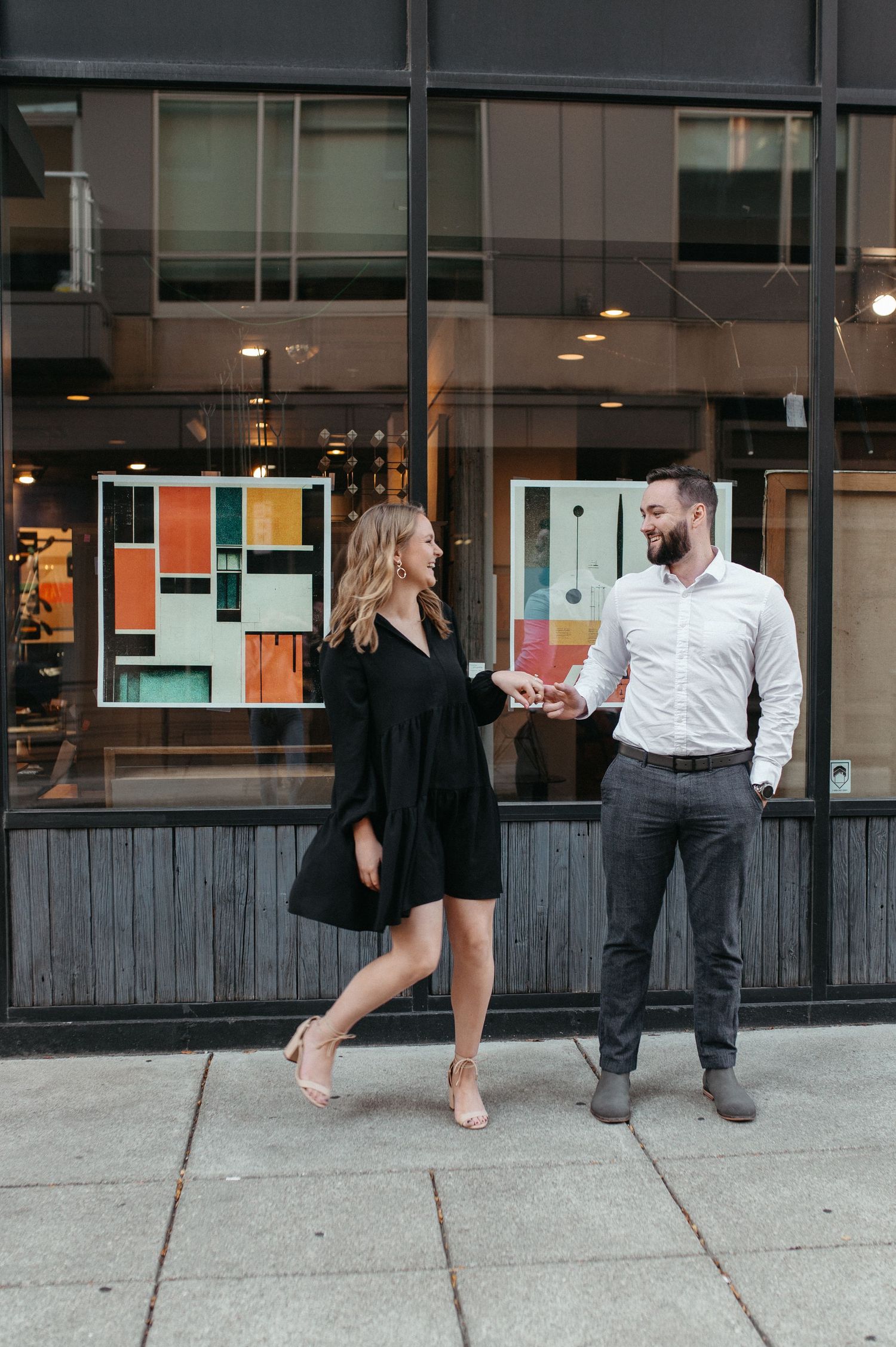 Playful couple dancing on sidewalk in front of modern building with colorful window display.