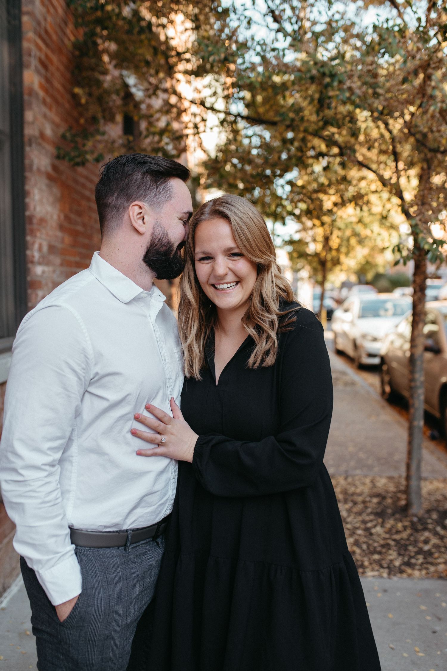 A romantic couple's portrait on a tree-lined street with fallen leaves and brick buildings.