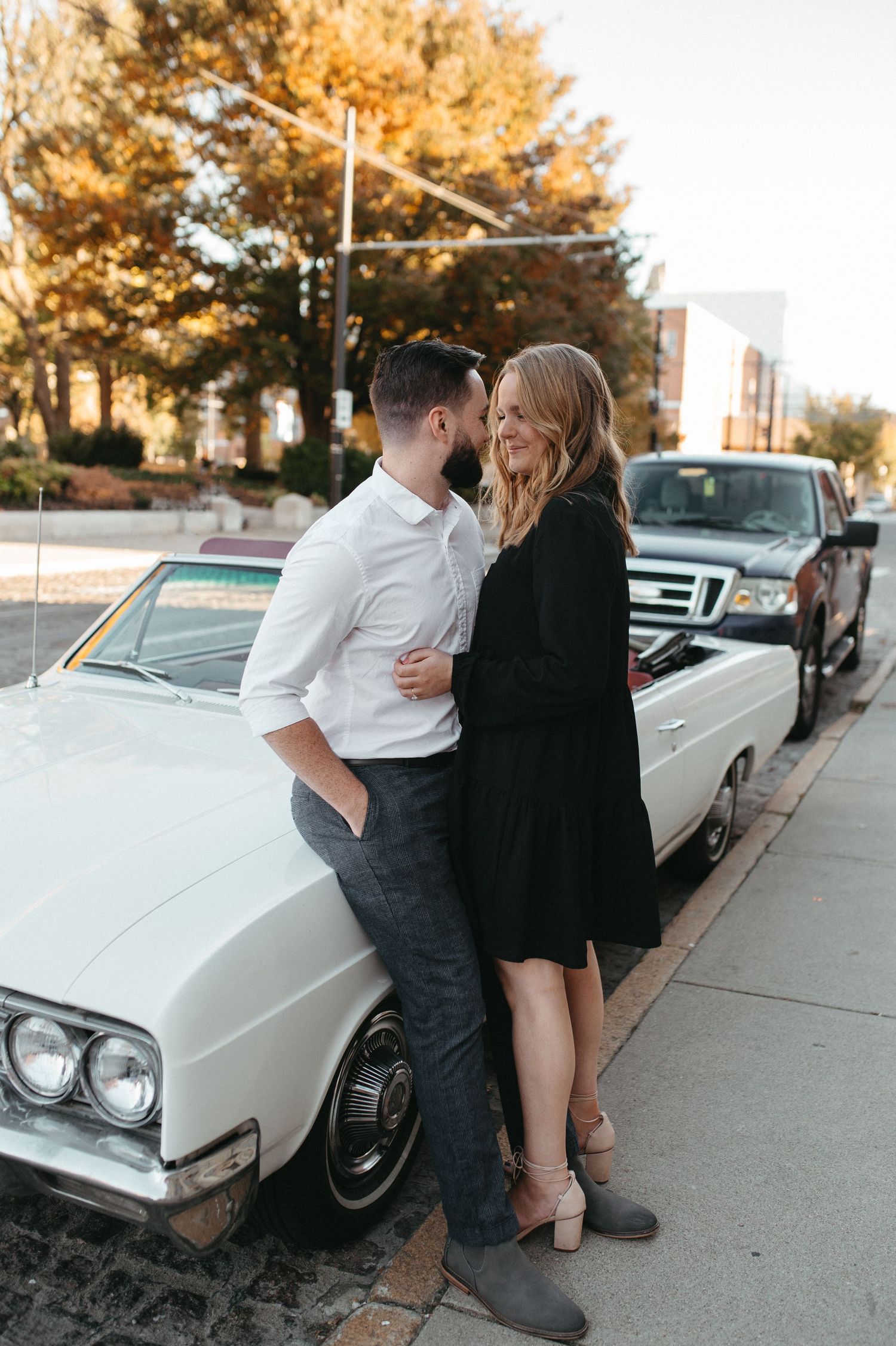 A romantic couple embraces beside a classic white car on an autumn street.
