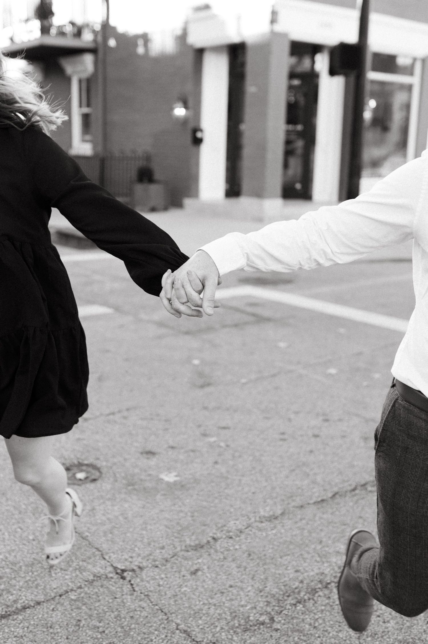 A black and white photo of two people holding hands, shot from the waist down on a city sidewalk.