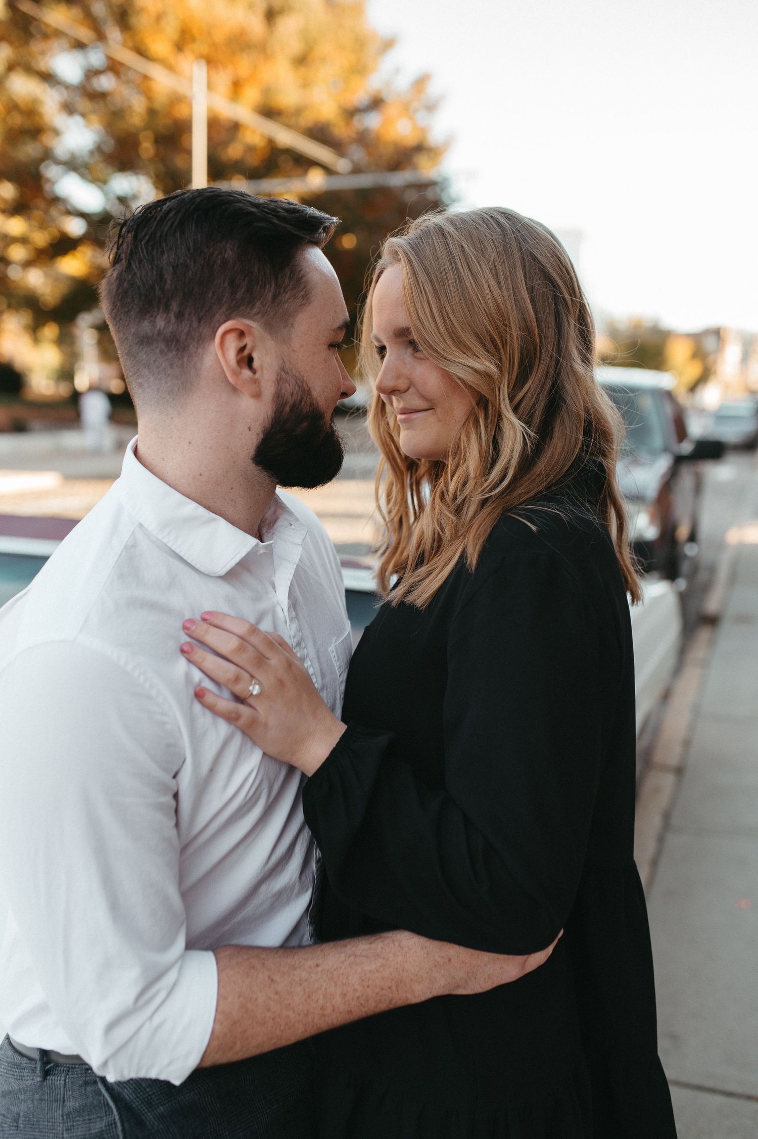 An engaged couple embraces in front of a vintage car during golden hour.