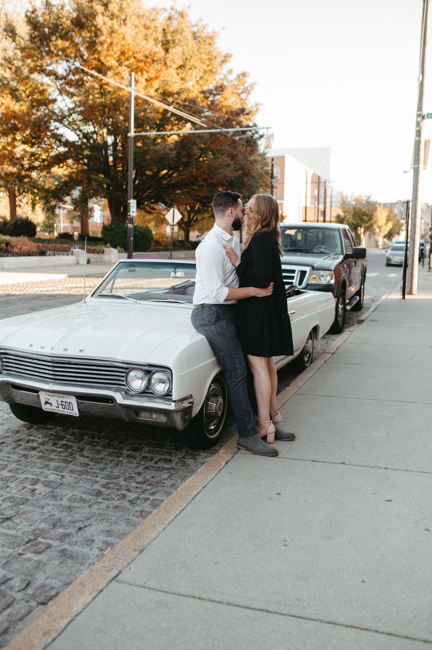 Couple poses romantically against a white vintage car on an autumn street.