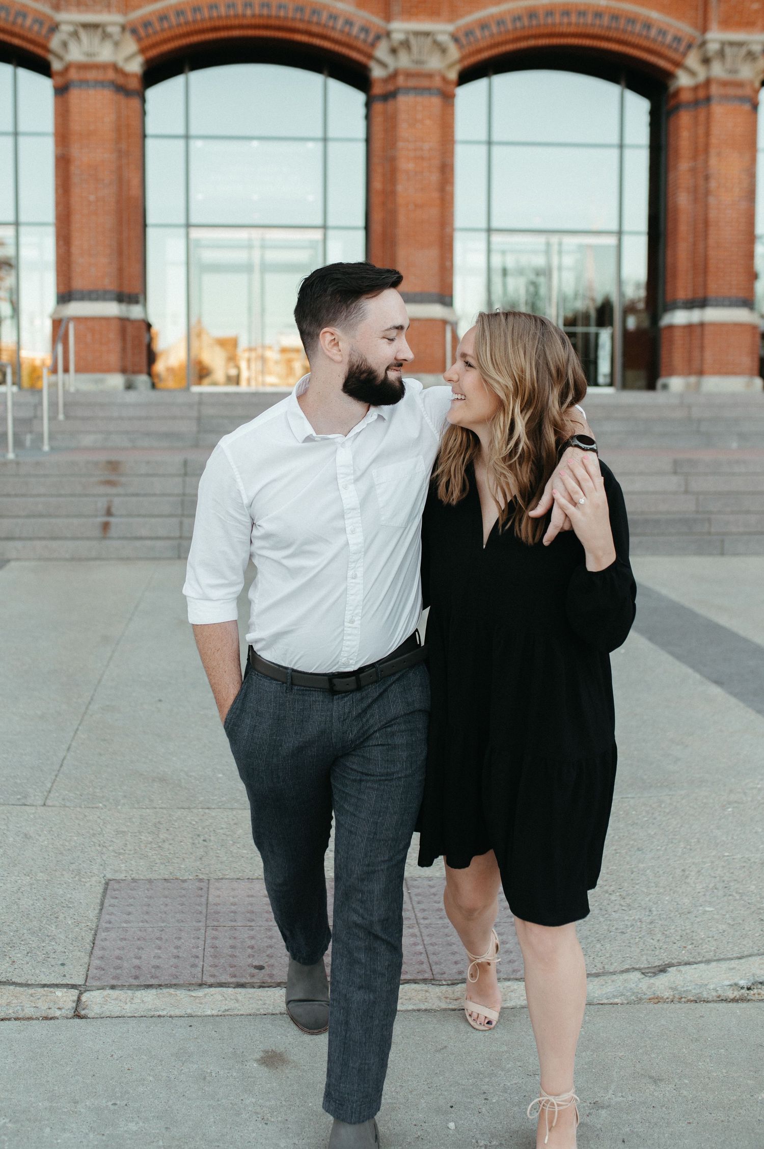 A stylish couple stands together in front of a brick building with arched windows.