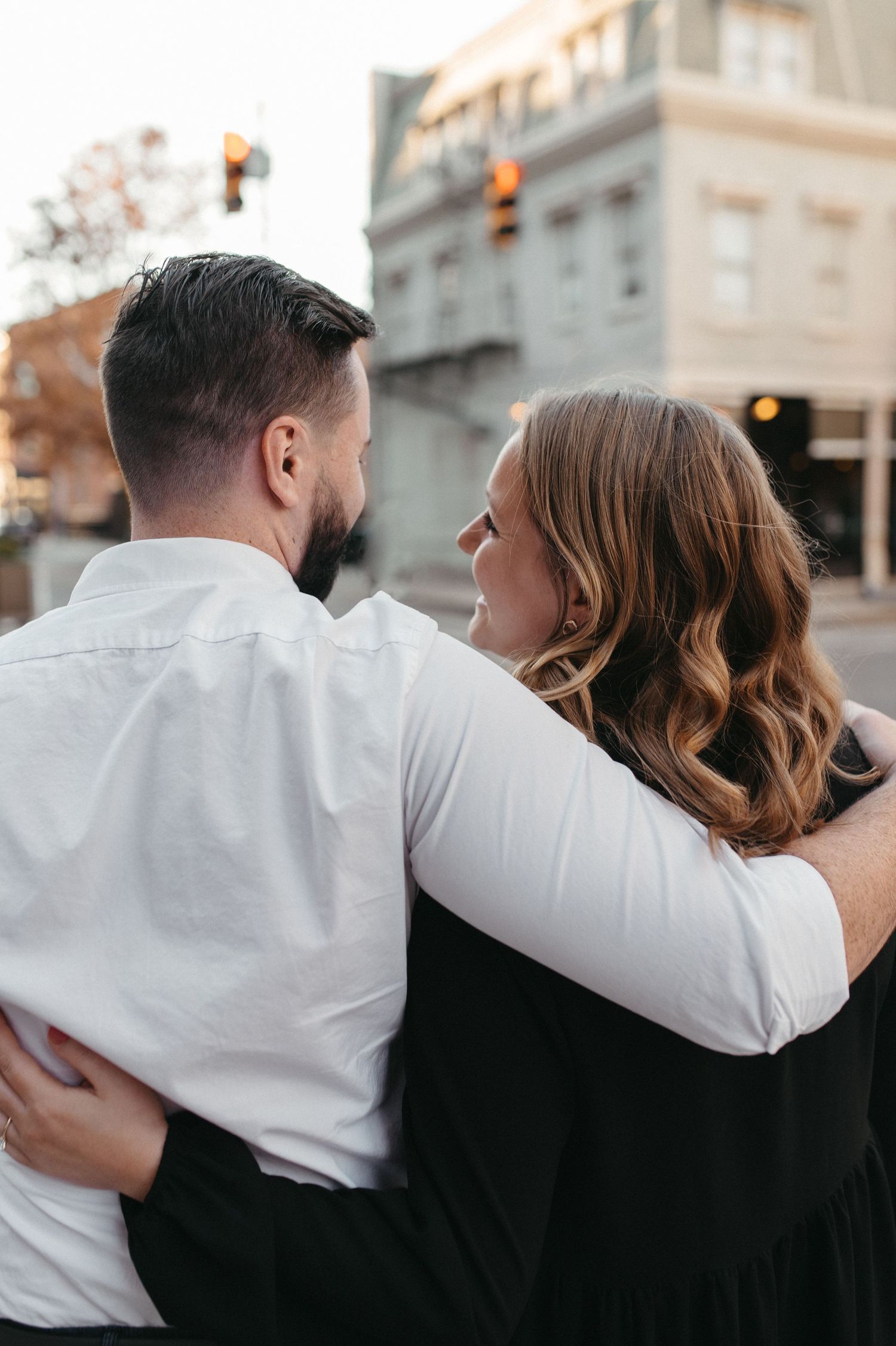 Couple in formal attire embracing on city sidewalk with historic architecture behind them.